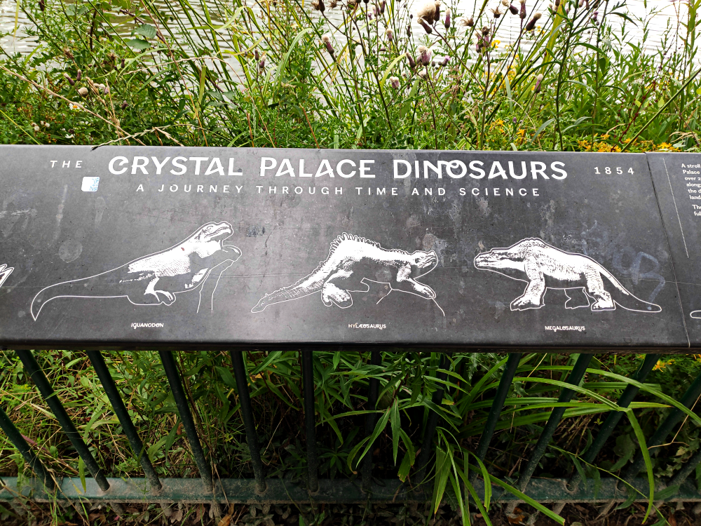 A photograph of an informational sign in London's Crystal Palace Park. The text on it reads, "The Crystal Palace Dinosaurs 1854, a journey through time and science". Three of the iconic Victorian dinosaur statures are also pictured below the title, showing the Iguanodon, Hylaeosaurus, and Megalosaurus.