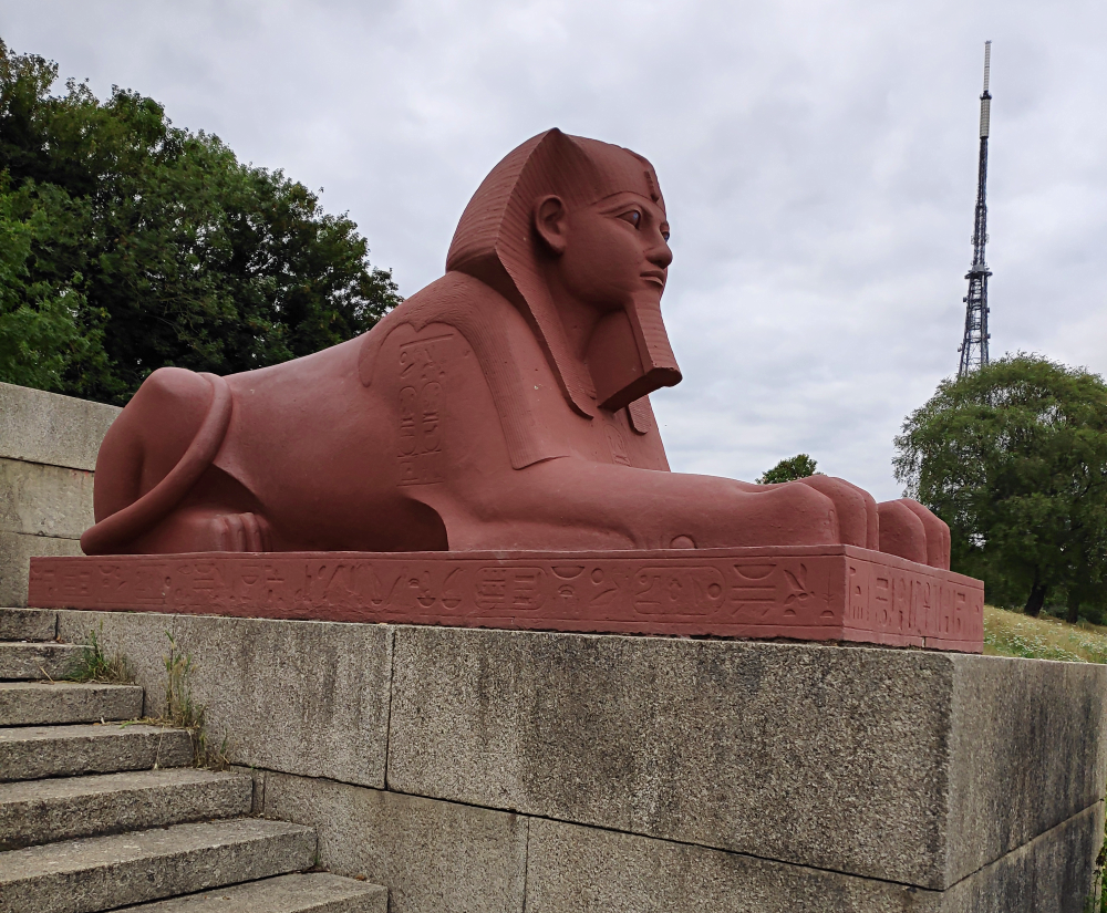 A photograph of one of the surviving sphinx statues in the Crystal Palace ruins, reclining on a plinth beside some stone steps. It's recently renovated with a coat of terracotta red paint to match its original Victorian-era appearance. In the background the huge Arqiva Crystal Palace telecom tower can be seen.