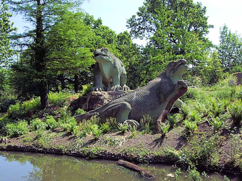 A clearer photograph of the two Crystal Palace Iguanodon, large bulky quadrupedal reptiles with beaked snouts, nose horns, four thick upright legs, scaly skin, and dragging tails. One is standing up and the other is reclining with one paw raised to rest on a cycad trunk.
