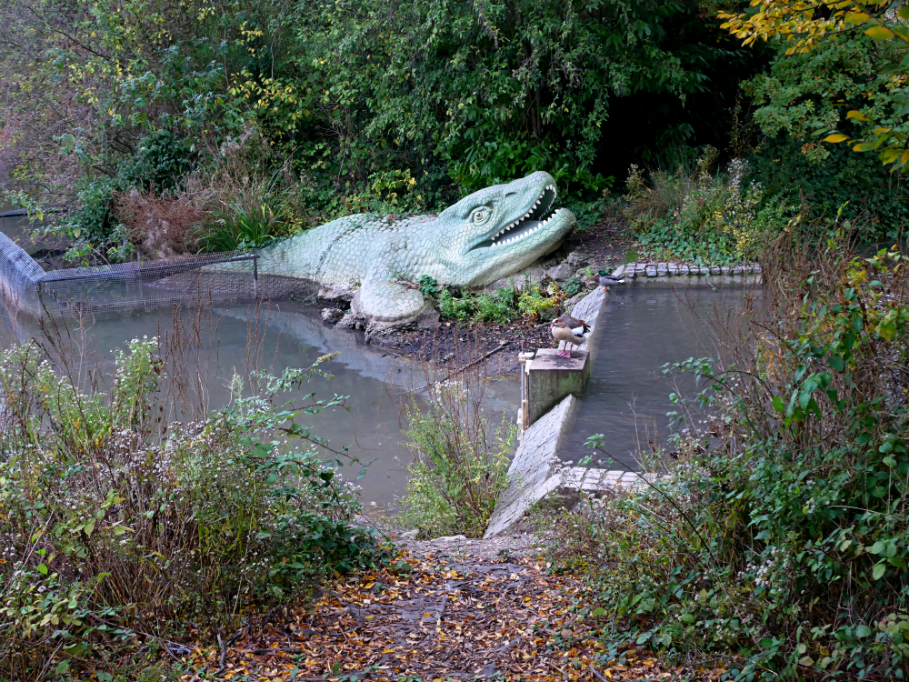 A photograph of the Crystal Palace mosasaur, an amphibious lizard-like animal lurking at the water's edge.