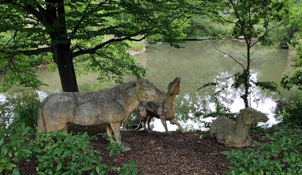 A photograph of the three Crystal Palace Anoplotherium statues. They're dog-like animals with camel-like heads, camel-like feet, and thick tails. One in the foreground on the left is in a standing pose, one in the midground on the right is in a reclining pose, and one int he background in the center is posed at the edge of a lake as if shaking water off itself.