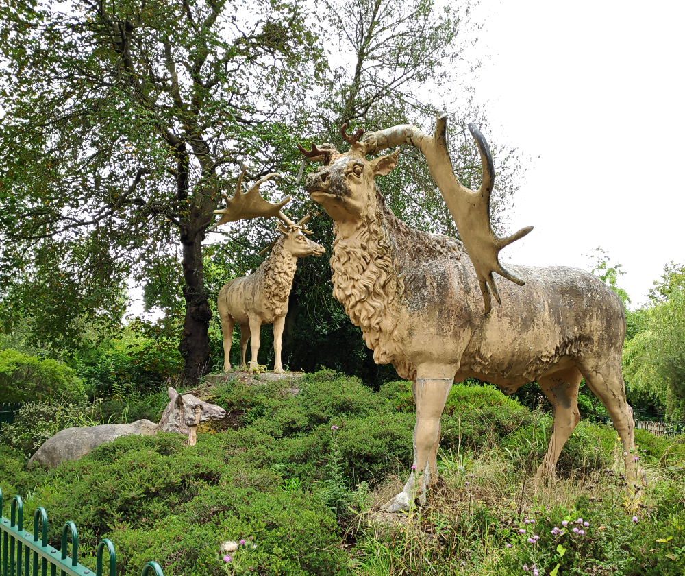 A photograph of the three Crystal Palace Megaloceros sculptures. They're giant deer depicted as resembling modern wapiti, with thick neck manes as if in their winter coats. Two are standing stags with enormous antlers, while the third is a reclining doe.