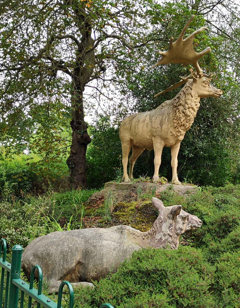A photograph of two of the Crystal Palace Megaloceros sculptures, a reclining doe and a standing stag. They're giant deer depicted as resembling modern wapiti, with thick neck manes as if in their winter coats.