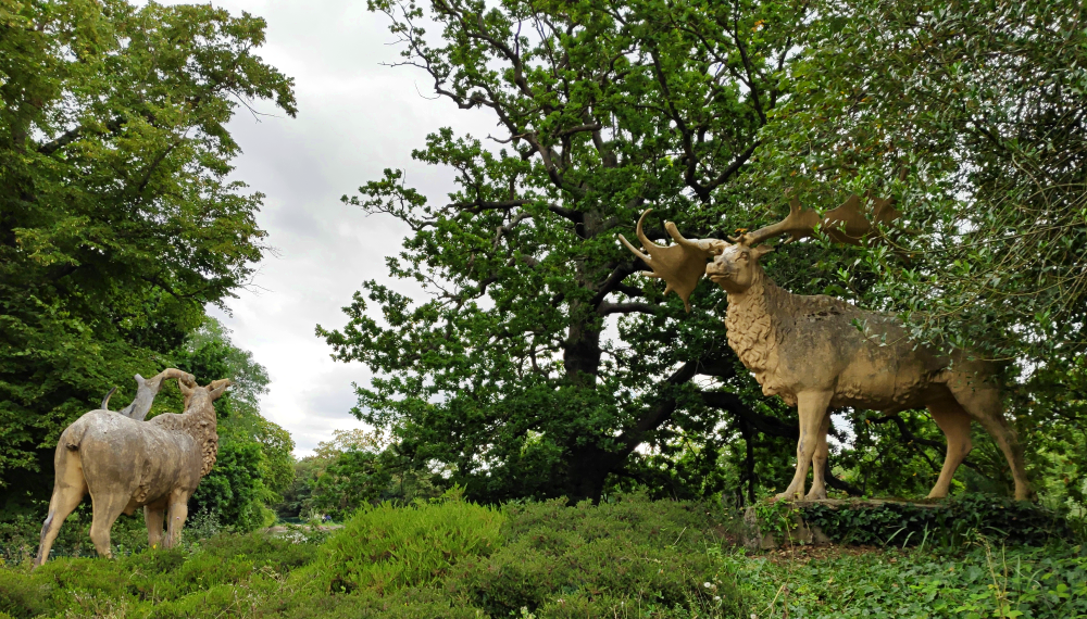 A photograph of two of the Crystal Palace Megaloceros sculptures, two standing stags. They're giant deer depicted as resembling modern wapiti, with thick neck manes as if in their winter coats. The left one is facing away from the viewer.