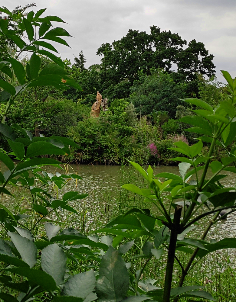 A photograph of the Crystal Palace Megatherium sculpture from the other side of the lake. Its head, upper body, and handless right arm can be distantly seen.